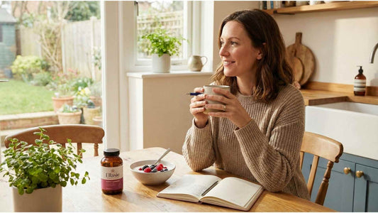 A relaxed woman journaling at a sunlit kitchen table with tea, a healthy breakfast, and a bottle of Ellasie women's probiotics, establishing a consistent daily wellness habit.