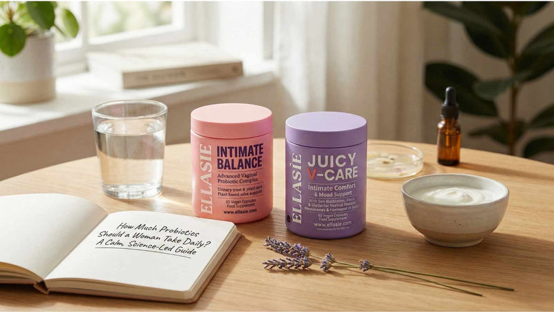 Ellasie Intimate Balance and Juicy V-Care probiotic supplements sitting on a wooden table next to a glass of water, a bowl of yogurt, and an open notebook
