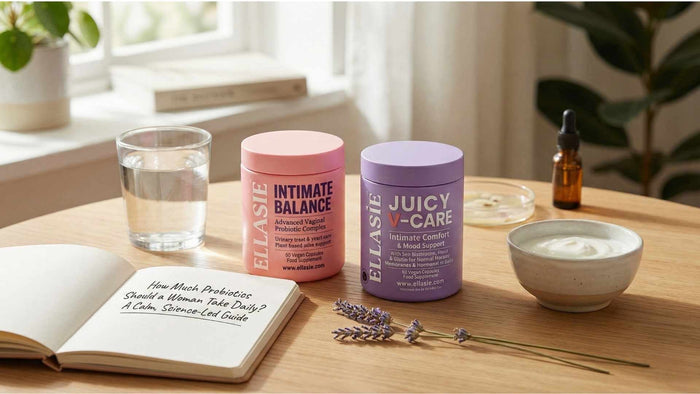 Ellasie Intimate Balance and Juicy V-Care probiotic supplements sitting on a wooden table next to a glass of water, a bowl of yogurt, and an open notebook
