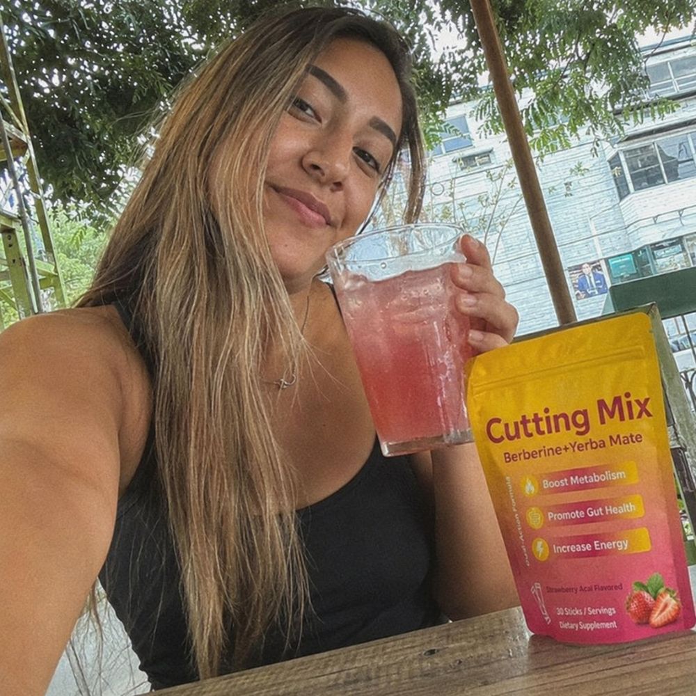 A woman holding a glass of pink iced drink next to a yellow and pink bag of Cutting Mix Berberine and Yerba Mate dietary supplement on a wooden table.