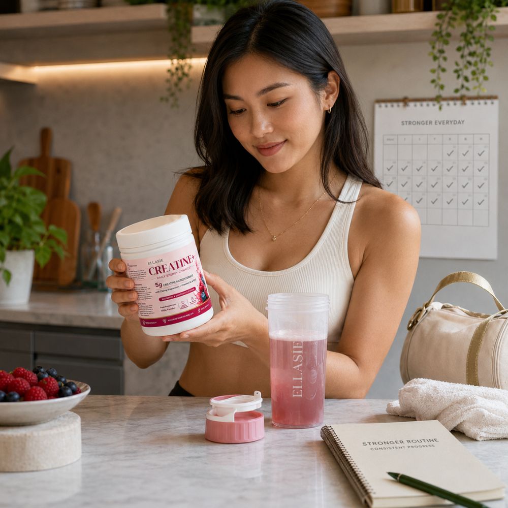 A woman in a kitchen setting holds a white tub of creatine powder labeled 'CREATINe' next to a pink and white travel water bottle. The scene includes a notebook, towel, and fruit on the counter.