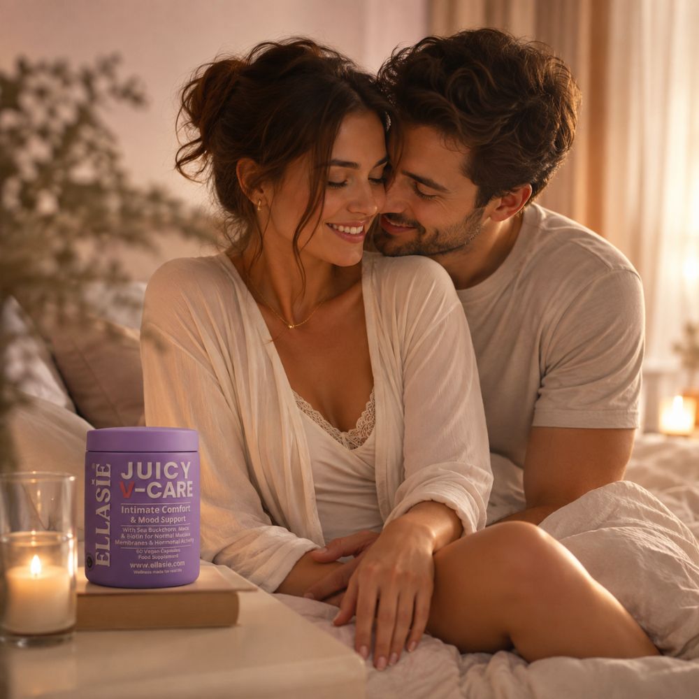 A couple sitting closely together on a bed in a warmly lit bedroom, with a purple tub of Eulasie Juicy V-Care on a small table in the foreground.