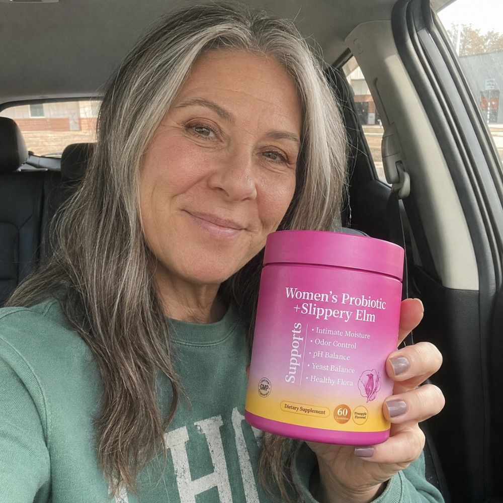 A woman with gray hair sitting in a car holds up a pink container of Women's Probiotic + Slippery Elm supplement. The container has a bright pink lid and body with a yellow gradient base, and the brand name 'Supports' is visible on the front.
