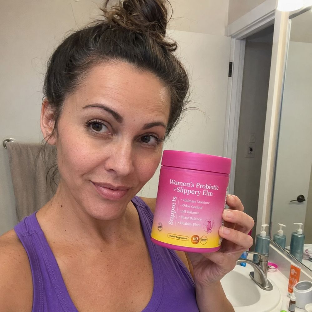 A woman in a purple tank top holds a jar of Women's Probiotic Slippary Elm in a bathroom setting.