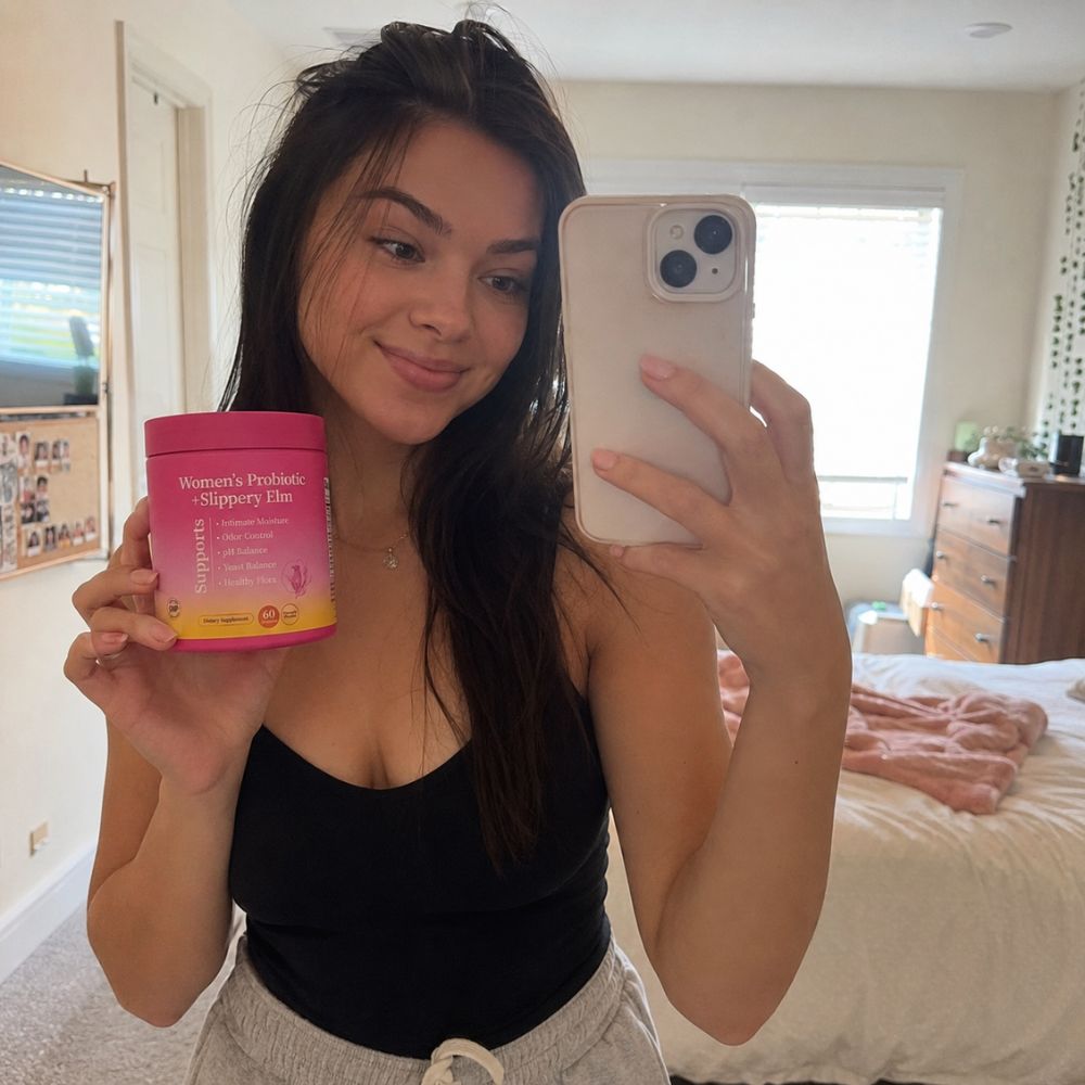 A woman taking a mirror selfie in a bedroom, holding a pink and yellow tub of Women's Probiotic Slurry Plus supplement. The tub has white text and a small pink graphic.