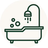 A dark green line icon of a bathtub with a shower head and bubbles, centered on a light beige circular background.