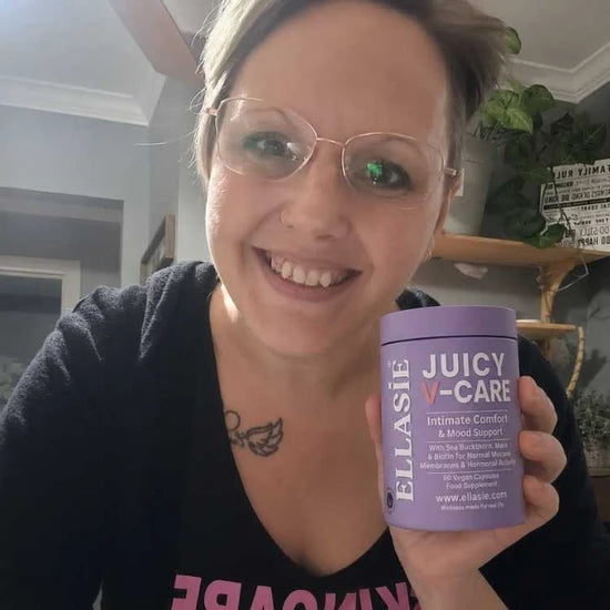A smiling woman with glasses holds up a purple tub of ELASIE Juicy V-Care intimate comfort and mood support cream in an indoor setting.