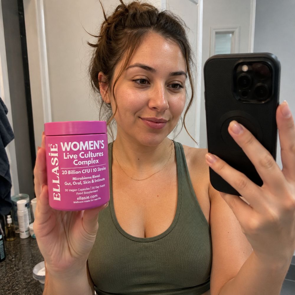 A woman taking a mirror selfie in a bathroom while holding a pink tub of ellasie Women's Live Cultures Complex supplement. The tub features white text and is shown next to her black smartphone.
