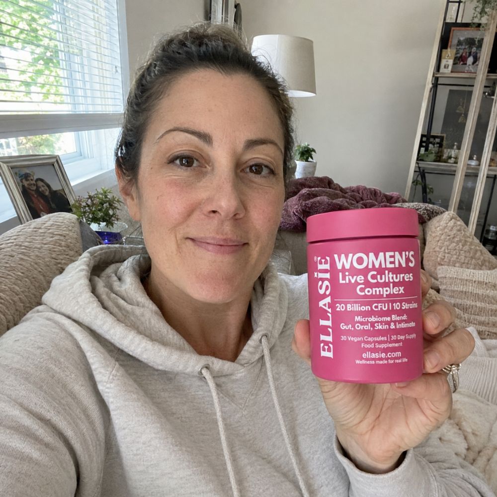 A woman in a light gray hoodie holds up a pink jar of Ellasie Women's Live Cultures Complex in front of a blurred home background.