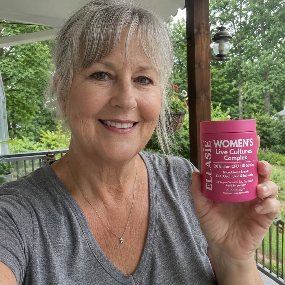 A smiling woman with short gray hair holds up a pink tub of ellasie Women's Live Cultures Complex' on a porch with green trees in the background.
