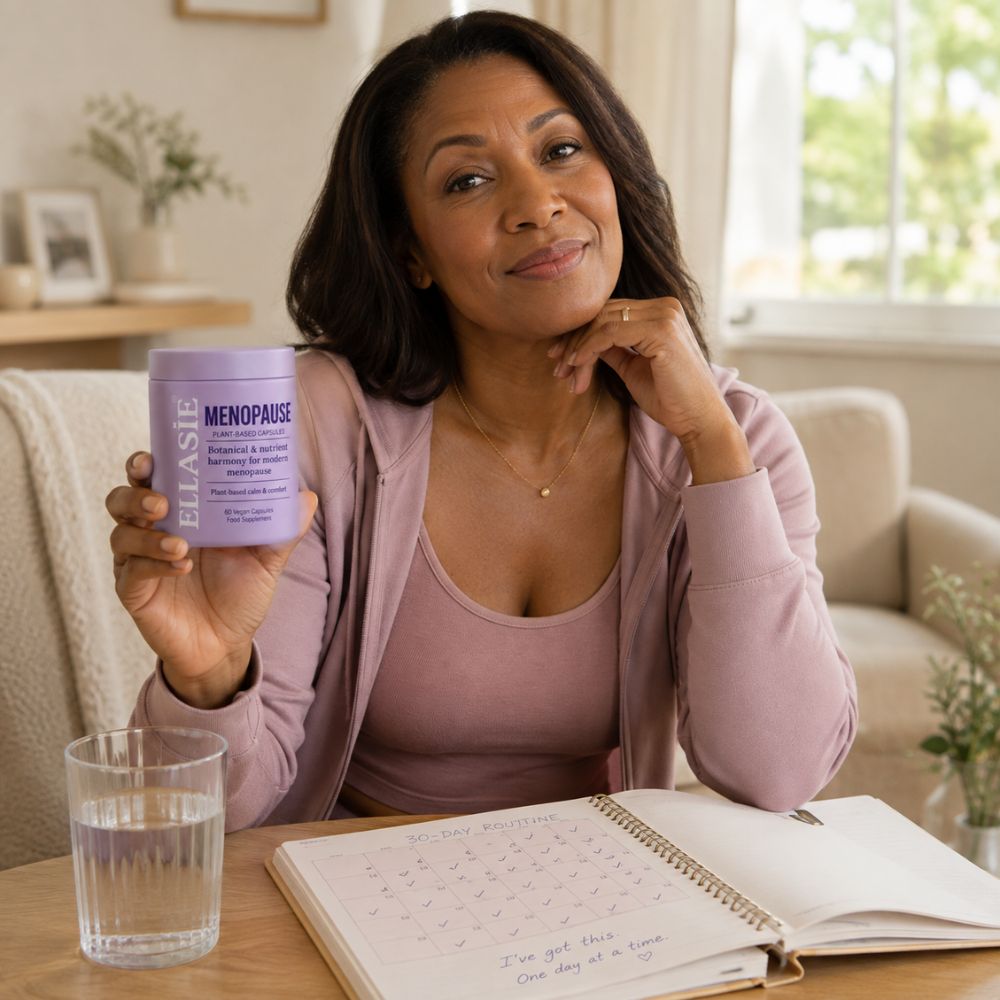 A woman sitting at a wooden table in a bright living room setting, holding up a lavender jar of ellasie Menopause Support capsules. An open notebook and a glass of water are on the table in front of her.