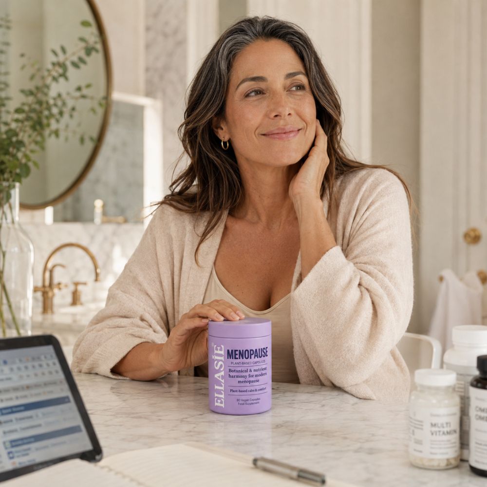 A woman in a beige cardigan sits at a marble counter holding a lavender jar labeled 'ELLASIE MENOPAUSE'. The setting is a bright, modern bathroom with gold fixtures, a mirror, and other supplement bottles on the counter.