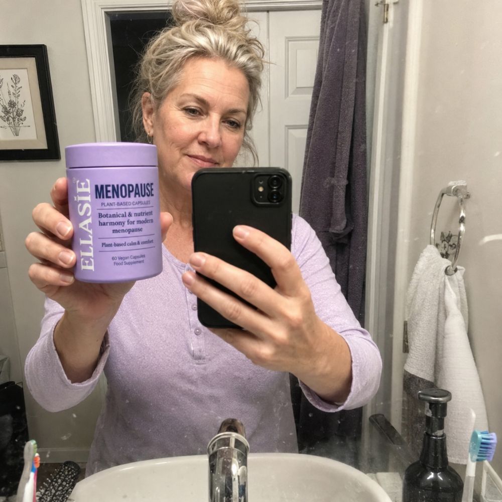 A woman taking a mirror selfie in a bathroom, holding up a lavender tub of ELLASIE Menopause Herbal & Symptomatic Relief Tea Extracts.