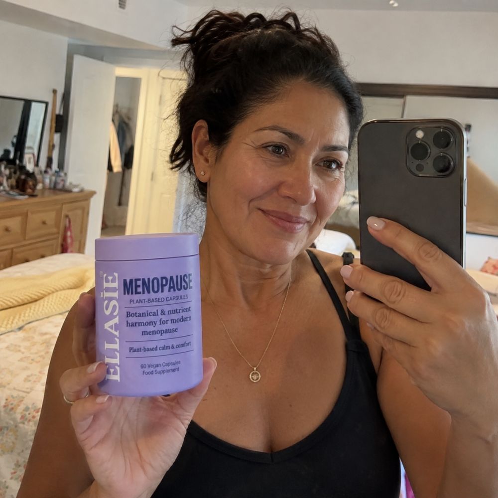 A woman taking a mirror selfie in a bedroom while holding a lavender jar of ELLASIE Menopause botanical and systemic health support cream next to her smartphone.