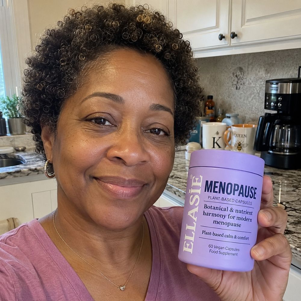 A smiling woman in a kitchen holds up a lavender purple tub of ELLASIE Menopause Plant Based Capsules. The tub is light purple with dark purple text, and the background shows white kitchen cabinets, a granite countertop, and a coffee maker.