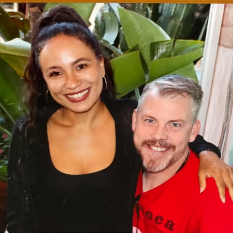 Two people standing together with green plants in the background