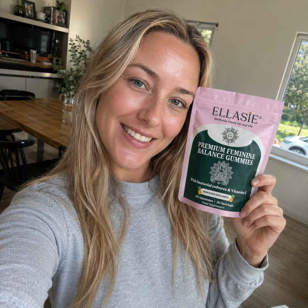 A woman holding a pink and green stand-up pouch of ELLASIE Premium Feminine Balance Gummies in a home setting.