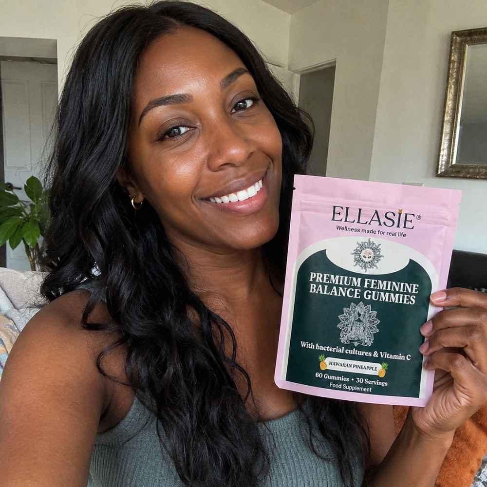 A smiling woman holds up a pink and dark green stand-up pouch of ELLASIE Premium Feminine Balance Gummies in a home setting.