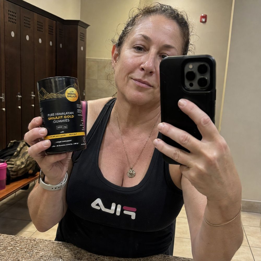 A woman taking a mirror selfie in a locker room, holding a black canister of 'Pure Himalayan Shilap Kripan Gulkor' alongside a black smartphone.