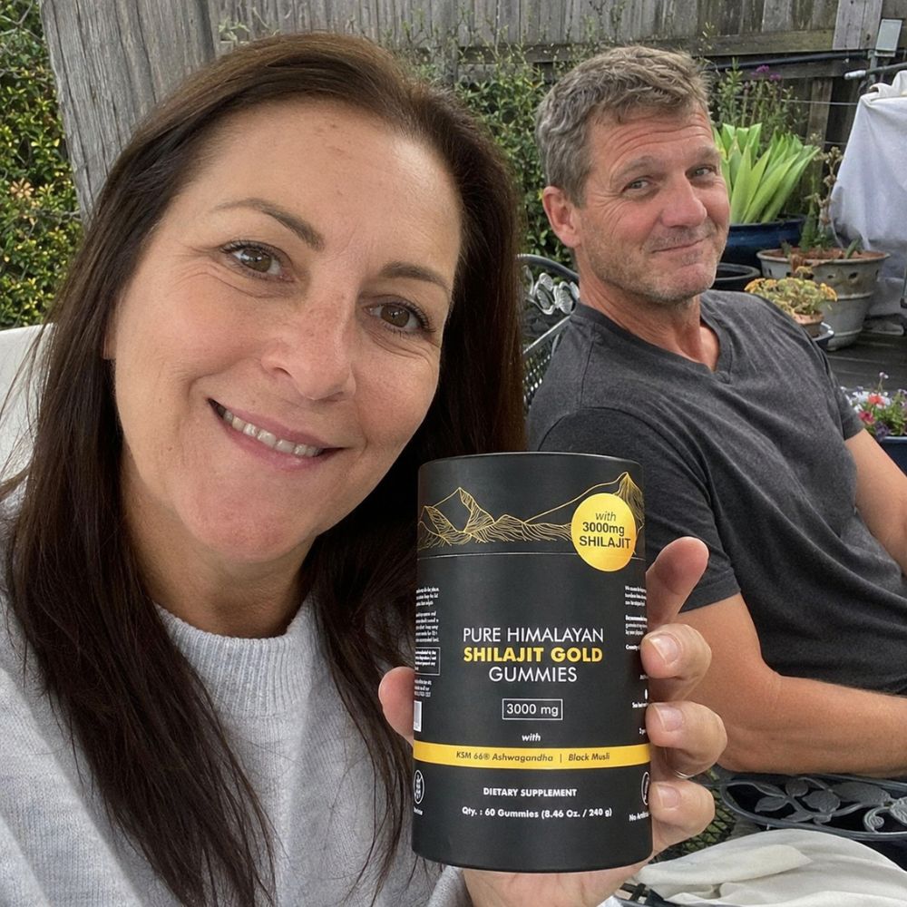 A woman holds a black canister of Shilajit Gold Gummies in front of her face, with a man sitting in a garden patio setting in the background.