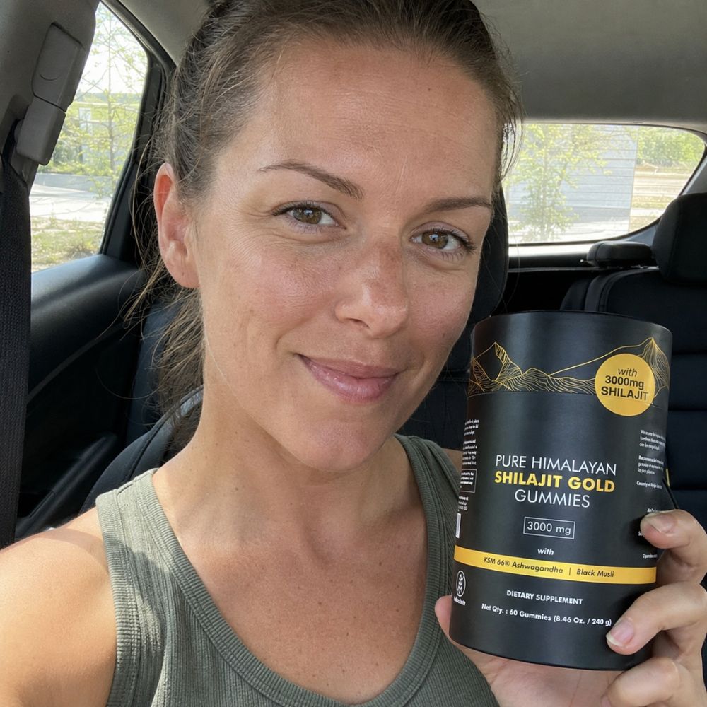 A woman sitting inside a car holds up a black container of Himalayan Shilajit Gold Gummies. The container is black with gold and white text, and a yellow circular badge that reads '100% DOING SHILAJIT'.
