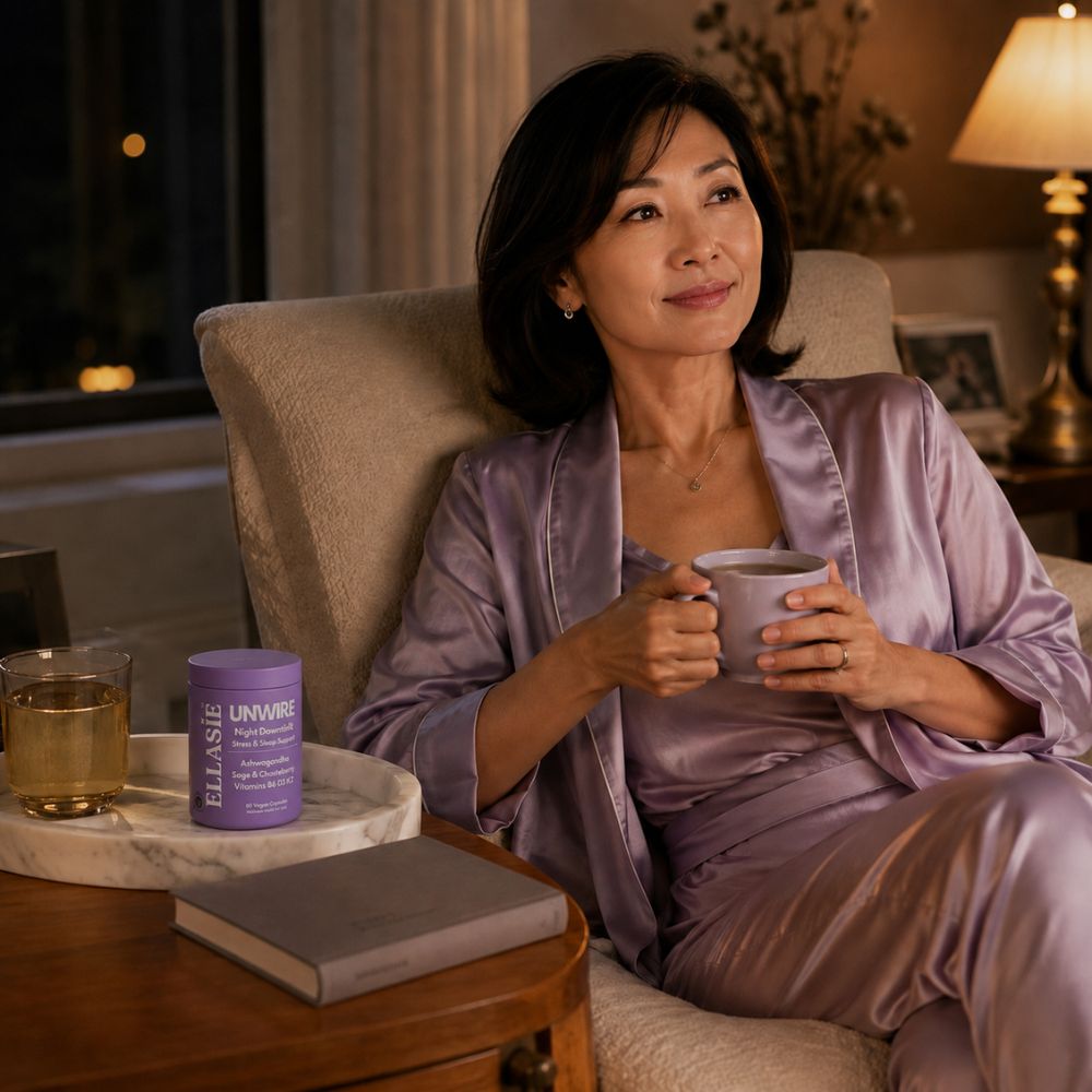 A woman in a lavender satin robe sits in a beige armchair holding a mug in a warmly lit living room. A purple cylindrical bottle labeled ellasie unwire pm rests on a marble tray beside her, with a book on a wooden table in the foreground.