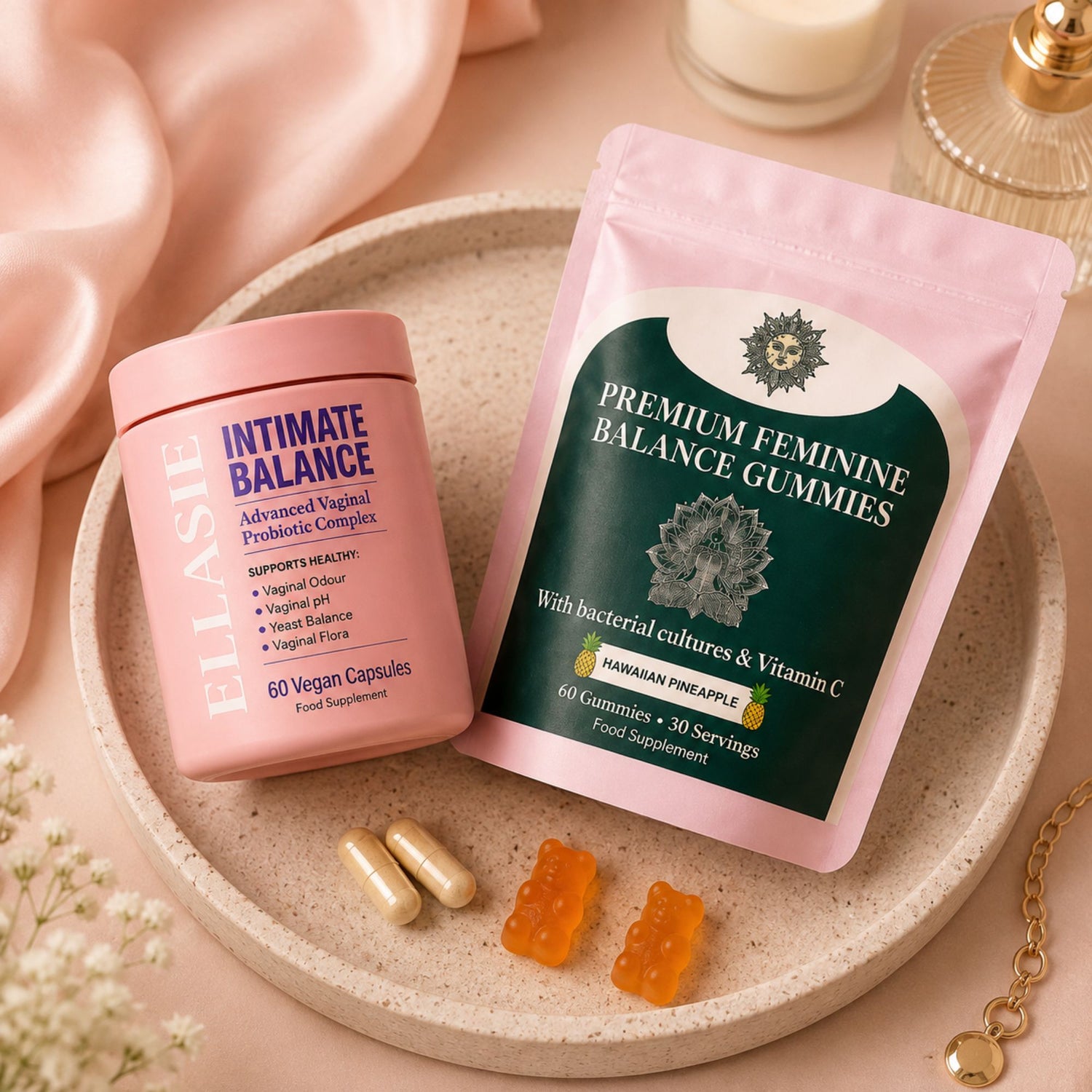 Two pink supplement products arranged on a round beige tray: a pink jar labeled 'Intimate Balance' and a pink stand-up pouch labeled 'Premium Feminine Balance Gummies.' The scene includes orange gummy bears, two beige capsules, soft pink fabric, and a softly blurred perfume bottle in the background.