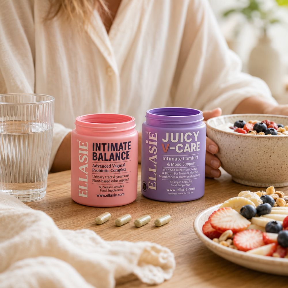 Two jars of ELLASIE probiotic supplements on a wooden table, one pink labeled 'Intimate Balance' and one purple labeled 'Juicy V-Care', with a person holding a bowl in the background and food plates nearby.