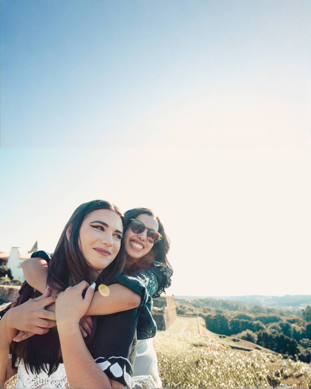 Two women hugging with a scenic background of trees and clear sky.